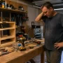 A cluttered home garage workspace with scattered tools, a half-finished wood project, and a man pausing mid-task while searching for a missing tool.